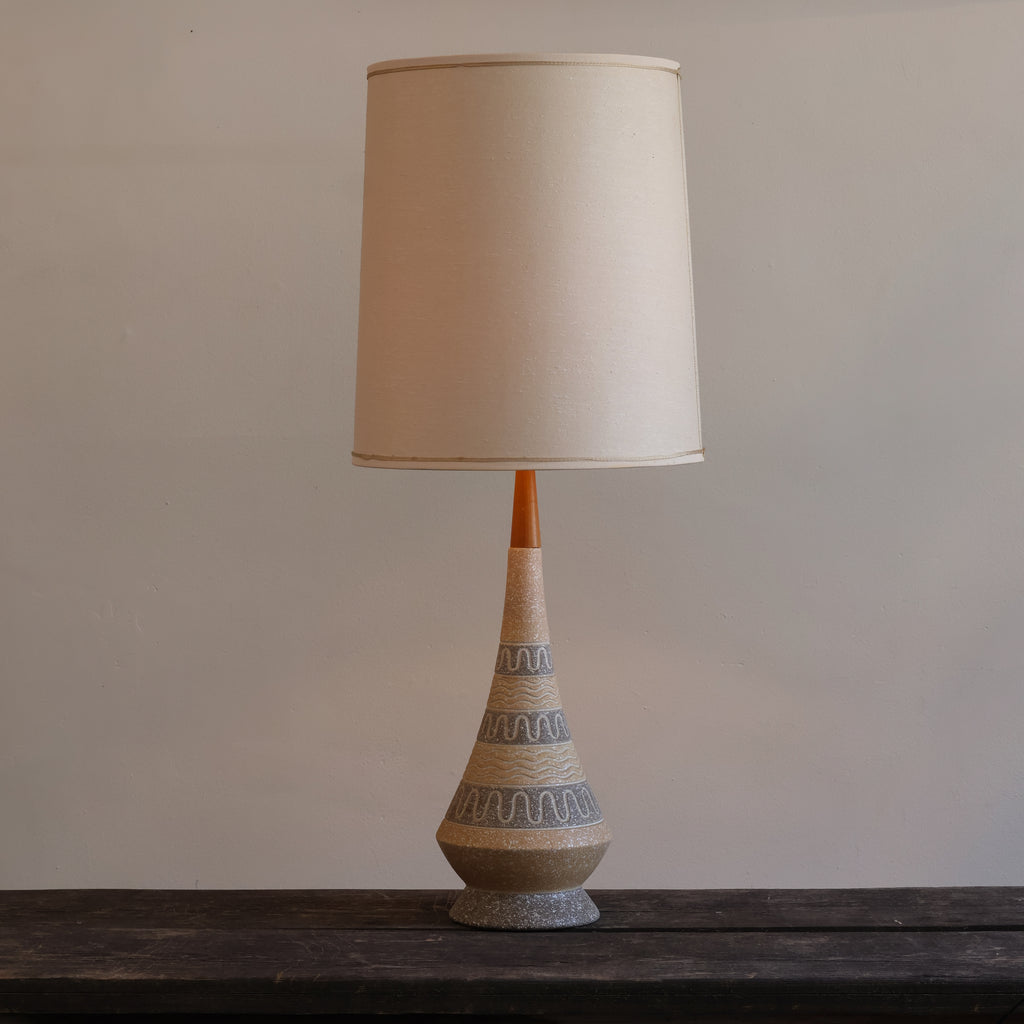 A mid-century modern table lamp with a tapered ceramic base featuring gray wavy patterns on a beige textured surface, topped with a warm orange neck and a large beige fabric drum shade.