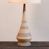 A mid-century modern table lamp with a textured, tapered ceramic base in beige and gray with wavy patterns, topped with a dark wood stem and a cream-colored fabric shade, set on a dark wooden surface against a neutral beige wall.