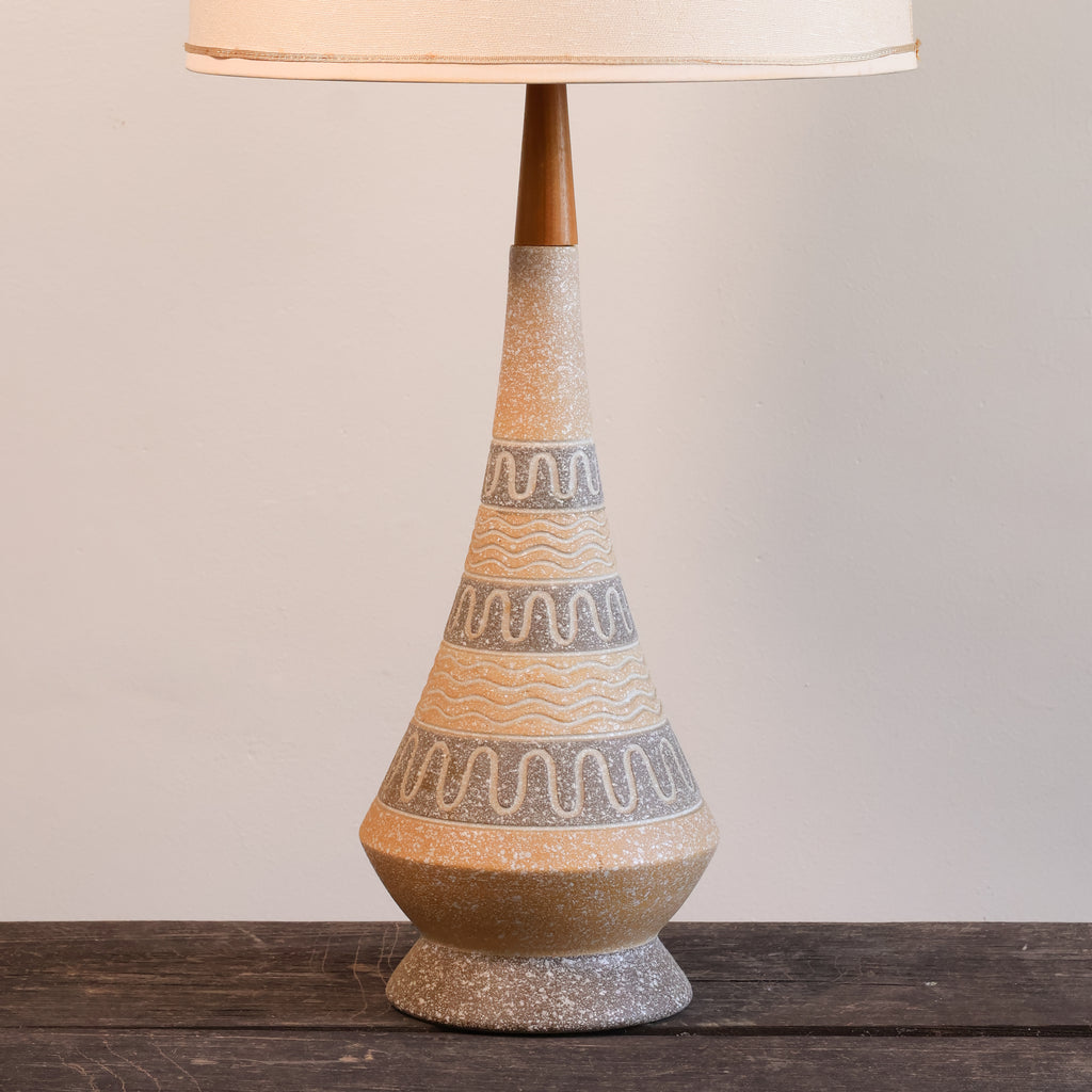 A mid-century modern table lamp with a textured, tapered ceramic base in beige and gray with wavy patterns, topped with a dark wood stem and a cream-colored fabric shade, set on a dark wooden surface against a neutral beige wall.