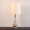 A table lamp with a tapered beige ceramic base featuring textured geometric patterns and a matching beige fabric drum shade, set on a dark wooden surface against a plain light gray wall.