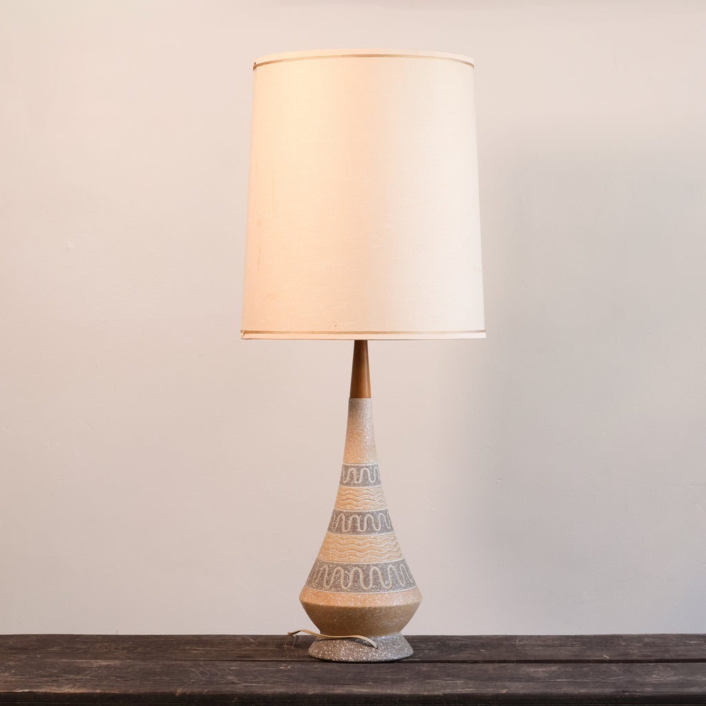 A table lamp with a tapered beige ceramic base featuring textured geometric patterns and a matching beige fabric drum shade, set on a dark wooden surface against a plain light gray wall.