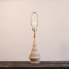 A mid-century modern table lamp with a tapered ceramic base, wrapped in beige and gray patterned fabric, topped with a warm brown wooden neck and a brass-colored metal shade frame holding a white bulb.