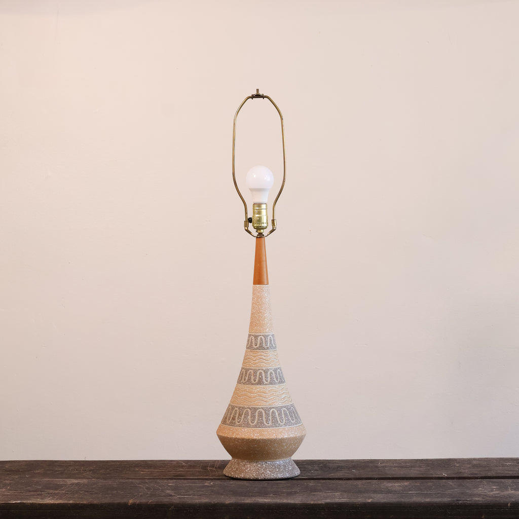 A mid-century modern table lamp with a tapered ceramic base, wrapped in beige and gray patterned fabric, topped with a warm brown wooden neck and a brass-colored metal shade frame holding a white bulb.