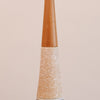 A close-up of a decorative lamp post or finial with a tapered wooden top and a glittery gold glitter cone base, set against a solid pale pink background.