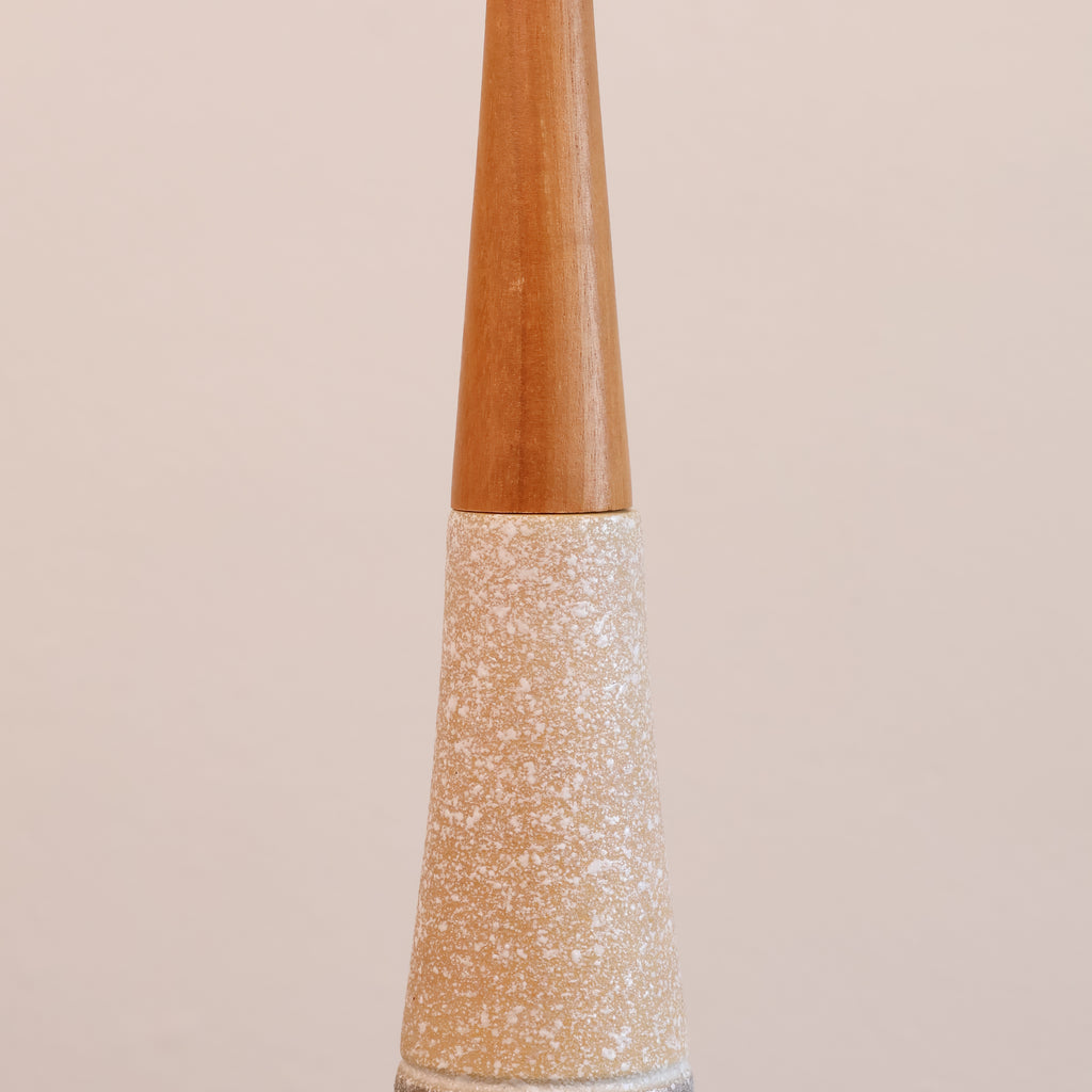 A close-up of a decorative lamp post or finial with a tapered wooden top and a glittery gold glitter cone base, set against a solid pale pink background.