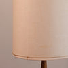 Close-up of a beige fabric lampshade with a visible seam and a thin metallic trim near the bottom edge, attached to a dark brown lamp base against a plain beige background.