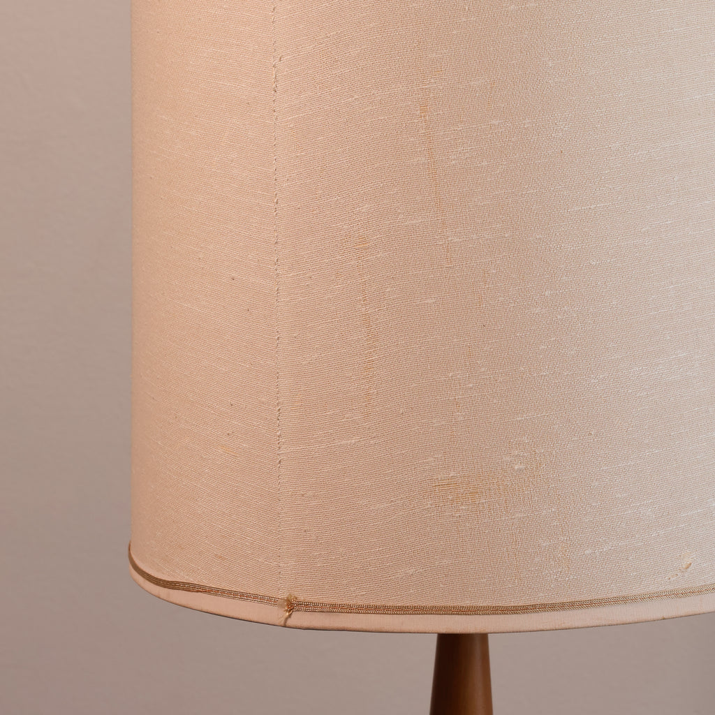 Close-up of a beige fabric lampshade with a visible seam and a thin metallic trim near the bottom edge, attached to a dark brown lamp base against a plain beige background.