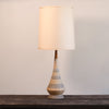 A table lamp with a tapered beige ceramic base featuring gray wavy patterns and a warm cream fabric drum shade, displayed on a dark wooden surface against a neutral light gray wall.