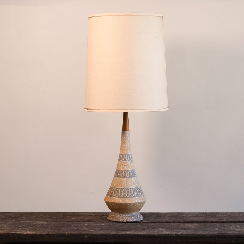A table lamp with a tapered beige ceramic base featuring gray wavy patterns and a warm cream fabric drum shade, displayed on a dark wooden surface against a neutral light gray wall.