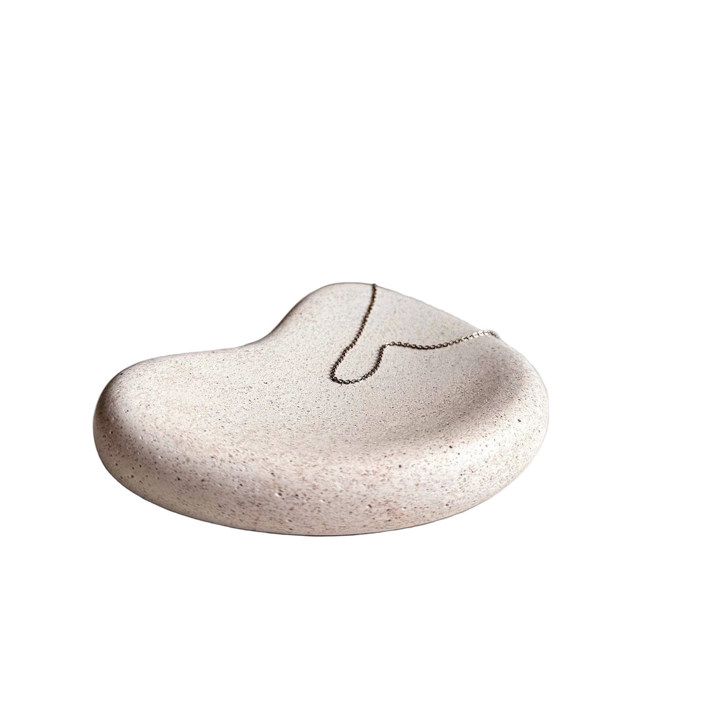 A small organic-shaped ceramic jewelry tray in a speckled cream-beige glaze, holding a delicate gold chain necklace, set on a white surface against a neutral beige background.