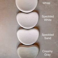 Four heart-shaped ceramic dishes in different finishes are stacked vertically on a textured beige surface. The labels next to each dish read 'White', 'Speckled White', 'Speckled Sand', and 'Creamy Gray'.