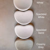 Four heart-shaped ceramic dishes in different finishes are stacked vertically on a textured beige surface. The labels next to each dish read 'White', 'Speckled White', 'Speckled Sand', and 'Creamy Gray'.