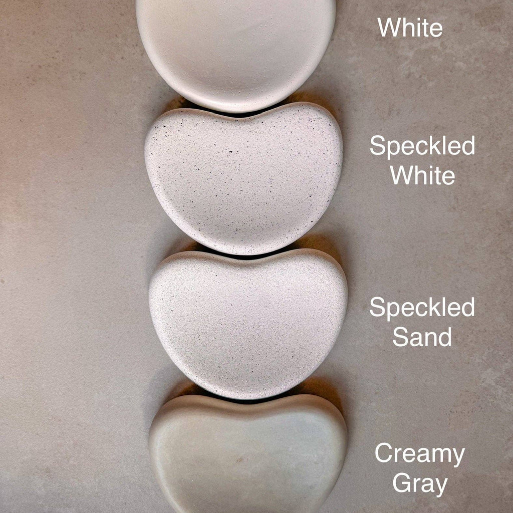 Four heart-shaped ceramic dishes in different finishes are stacked vertically on a textured beige surface. The labels next to each dish read 'White', 'Speckled White', 'Speckled Sand', and 'Creamy Gray'.