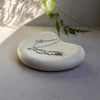 A delicate silver chain necklace with a small pendant rests on a smooth, rounded white stone dish. The setup is styled with soft shadows cast by hanging green ferns on a neutral gray background.