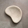 A small organic-shaped ceramic dish or soap dish in a light cream or off-white color, photographed on a textured gray surface.
