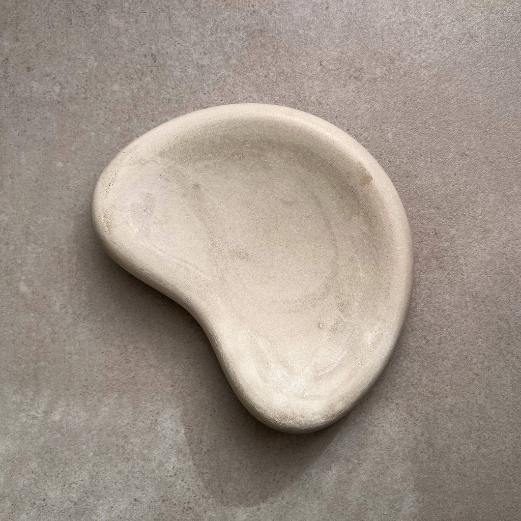 A small organic-shaped ceramic dish or soap dish in a light cream or off-white color, photographed on a textured gray surface.