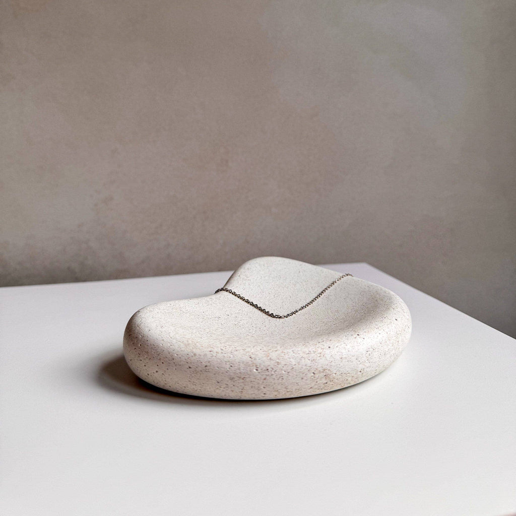 A small organic-shaped soap dish made of speckled cream stone, holding a delicate silver chain necklace, set on a white surface against a textured beige wall.