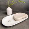 A minimalist bathroom set featuring a large oval white stone soap dish with a matching curved soap bar, a small matte white ceramic vase with green bamboo stems, and a small silver ring on a textured gray concrete surface.