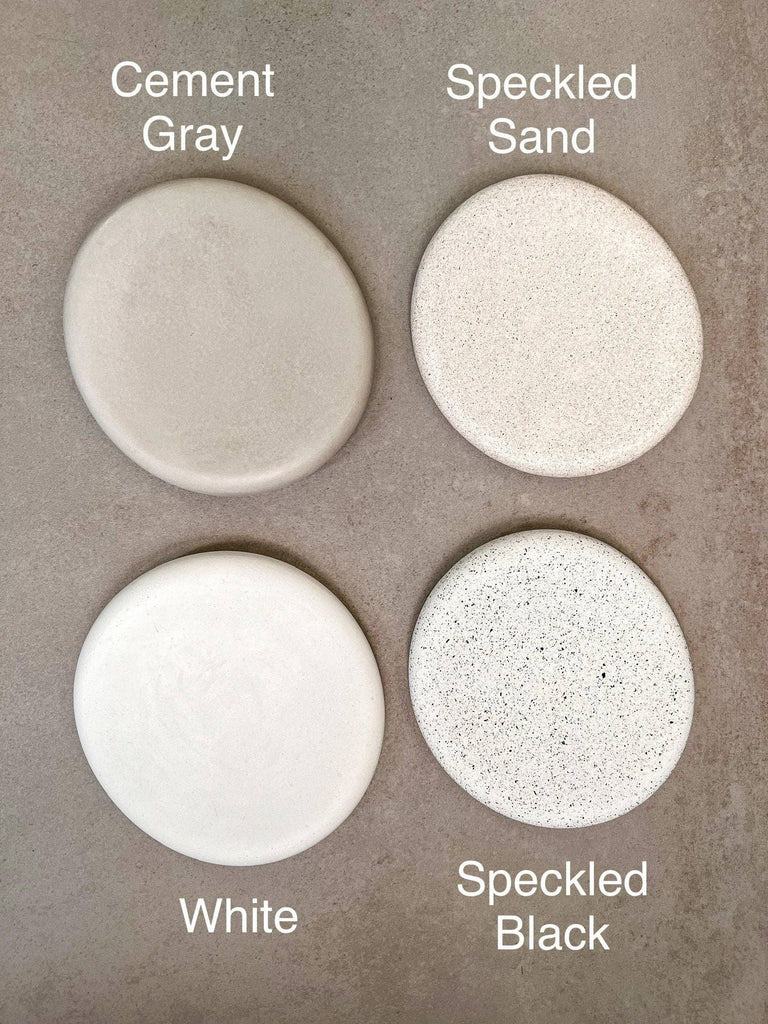 Four circular stone or concrete swatches arranged in a 2x2 grid on a textured taupe background, labeled Cement Gray, Speckled Sand, White, and Speckled Black.