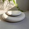 Two smooth, rounded stone-like decorative pieces stacked in a small set, photographed on a neutral surface with soft natural shadows and green leaves in the background.