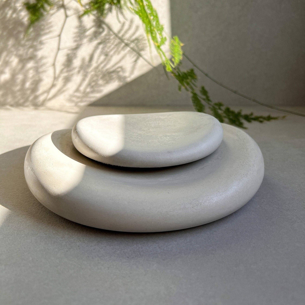 Two smooth, rounded stone-like decorative pieces stacked in a small set, photographed on a neutral surface with soft natural shadows and green leaves in the background.