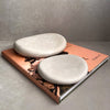 Two smooth, oval ceramic coasters in a light cream color, displayed on top of a peach-colored book against a textured gray background.