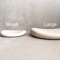 Two minimalist curved soap dishes in matte ceramic, labeled 'Small' and 'Large' to show size options, set against a neutral textured gray background.