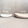 Two minimalist curved soap dishes in matte ceramic, labeled 'Small' and 'Large' to show size options, set against a neutral textured gray background.