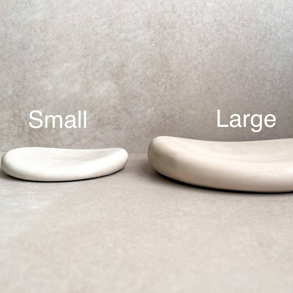 Two minimalist curved soap dishes in matte ceramic, labeled 'Small' and 'Large' to show size options, set against a neutral textured gray background.