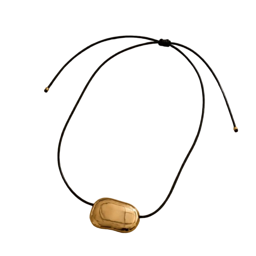 A gold-toned capsule pendant necklace on a black adjustable cord, displayed on a textured beige surface with soft diagonal shadows.