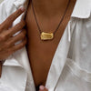 A close-up of a person wearing a gold abstract pendant necklace on a black cord, styled with an open white shirt.