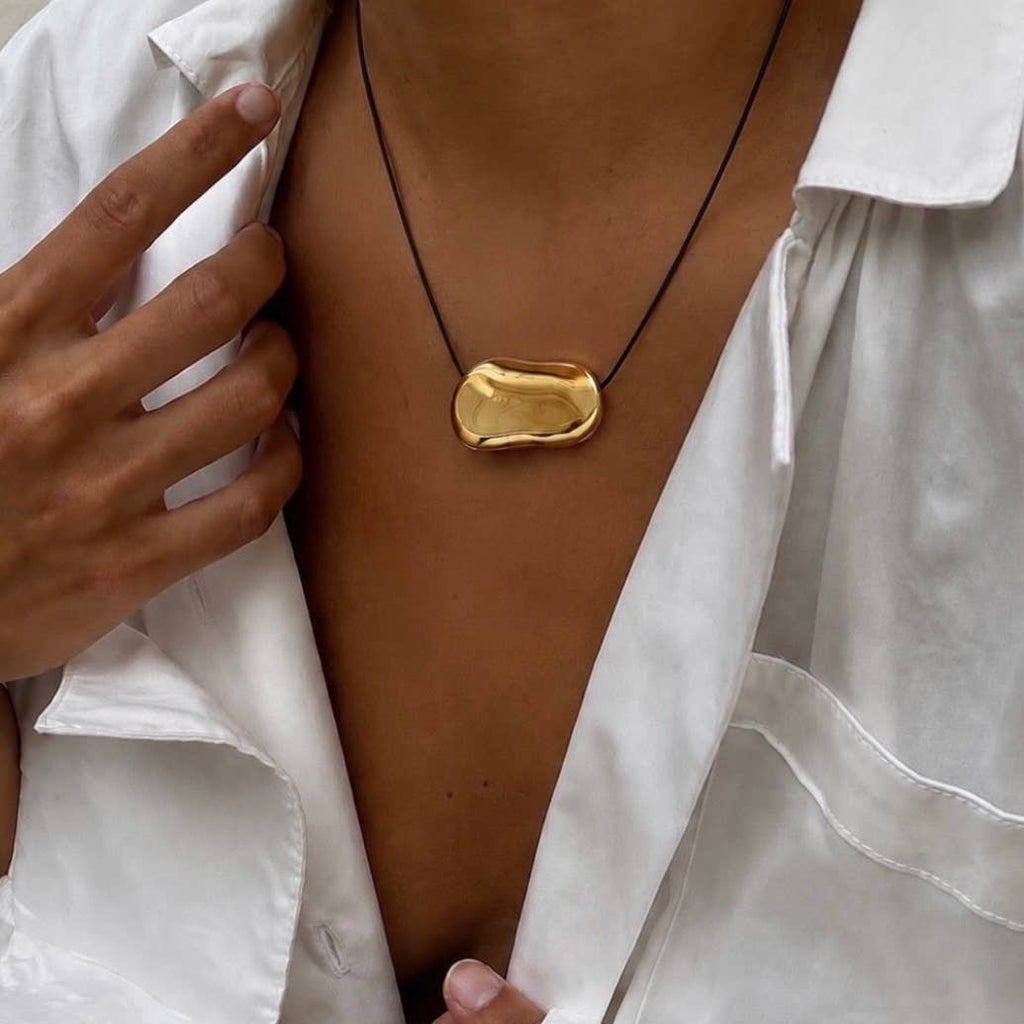 A close-up of a person wearing a gold abstract pendant necklace on a black cord, styled with an open white shirt.