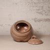 A rustic handmade ceramic jar with its lid resting beside it, featuring a rounded body, open top, and a small loop handle on the lid. The jar and lid are glazed in a warm, earthy brownish-beige glaze against a textured, distressed off-white wall.