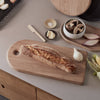 Wooden cutting board with bread on a kitchen counter with various items.