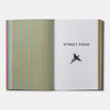 An open book or magazine on a light gray background. The left page has vertical stripes in green, pink, and blue, while the right page is white with the text 'STREET FOOD' and a black bird silhouette.