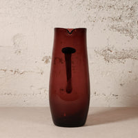 A tall, dark amber-brown translucent glass vase with a central cutout, standing on a light surface against a textured off-white wall.