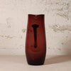 A tall, dark amber-brown translucent glass vase with a central cutout, standing on a light surface against a textured off-white wall.