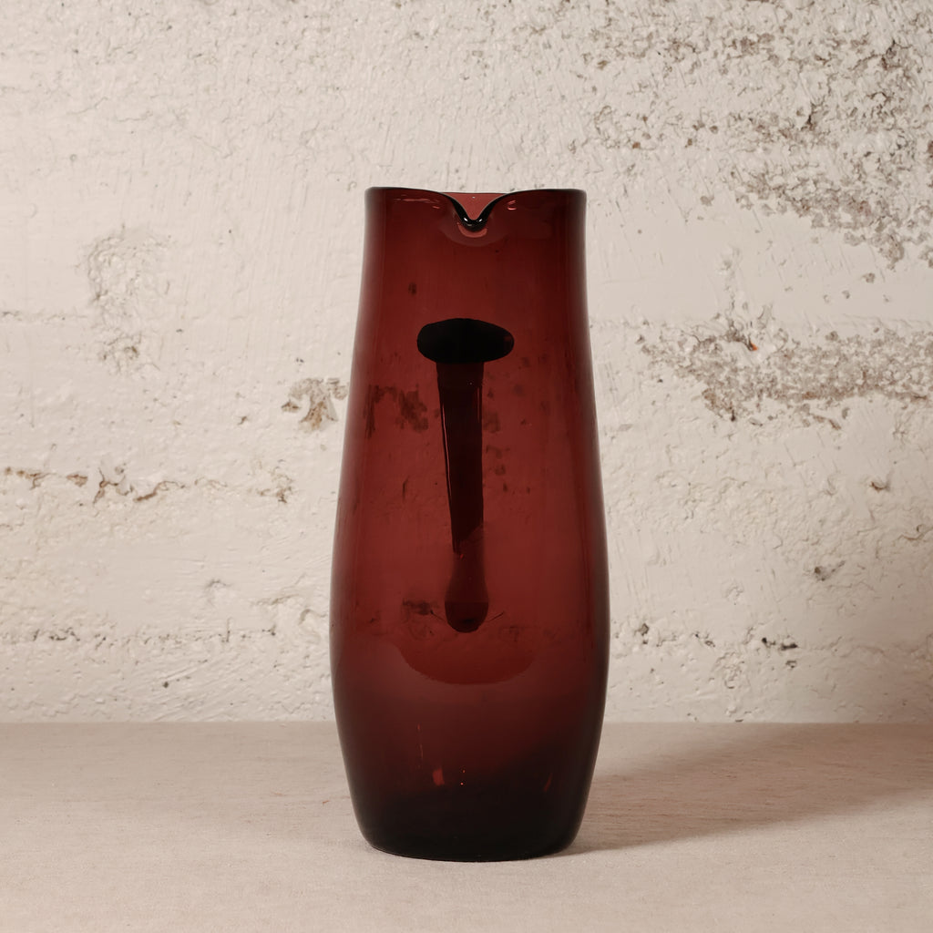 A tall, dark amber-brown translucent glass vase with a central cutout, standing on a light surface against a textured off-white wall.