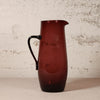 A translucent dark red glass pitcher with a black handle, standing on a light surface against a textured off-white wall.
