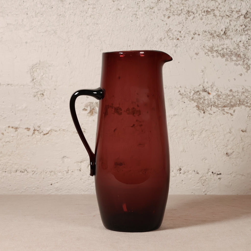 A translucent dark red glass pitcher with a black handle, standing on a light surface against a textured off-white wall.