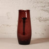 A tall, dark amber-brown translucent glass pitcher with a handle, standing on a light beige surface against a textured off-white wall.