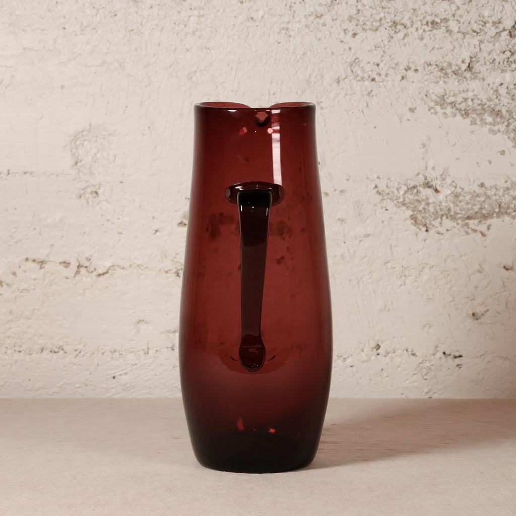 A tall, dark amber-brown translucent glass pitcher with a handle, standing on a light beige surface against a textured off-white wall.