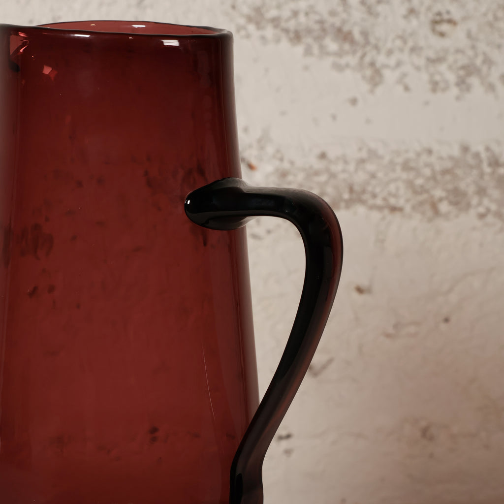 A close-up of a translucent dark red glass pitcher with a curved handle, set against a textured off-white wall.