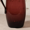 A close-up of a translucent dark red glass pitcher with a handle, resting on a light beige textured surface against a white brick wall.