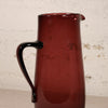 A translucent dark red glass pitcher with a curved black handle, shown against a textured off-white brick wall background.