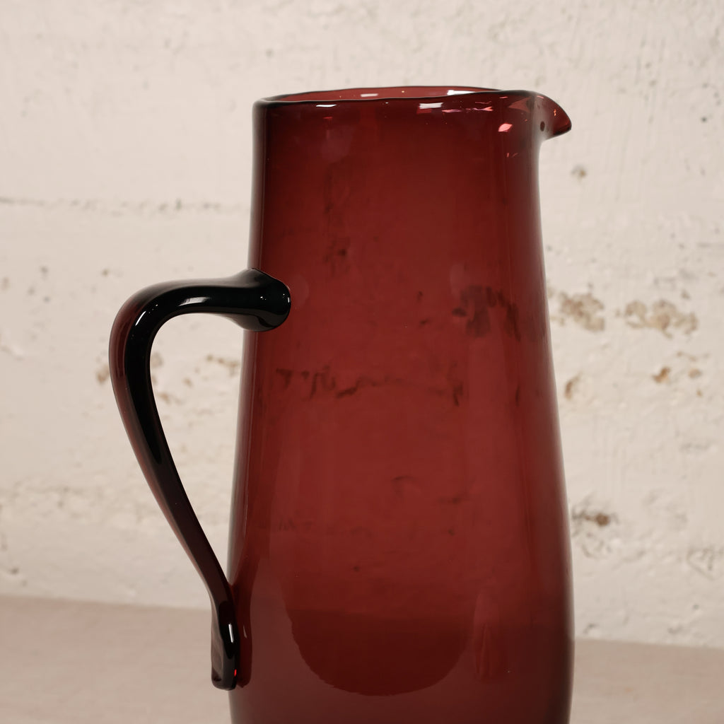 A translucent dark red glass pitcher with a curved black handle, shown against a textured off-white brick wall background.