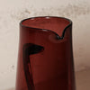 A close-up of a translucent dark red glass pitcher with a small pouring spout, set against a textured off-white wall.