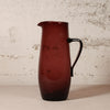 A dark amber-brown translucent glass pitcher with a black handle, standing on a light surface against a textured off-white wall.