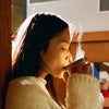 Person drinking from a mug with steam, wearing a light-colored sweater indoors.