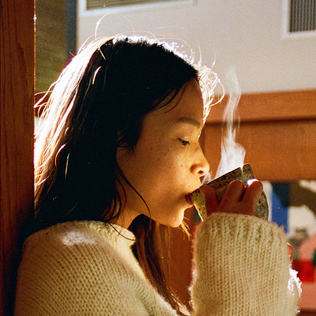 Person drinking from a mug with steam, wearing a light-colored sweater indoors.